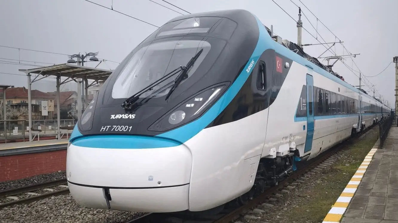 A white and turquoise TURASAS-branded high-speed train unit numbered HT 70001 stands at a platform on a grey overcast day, with overhead electrification cables visible and residential buildings in the background