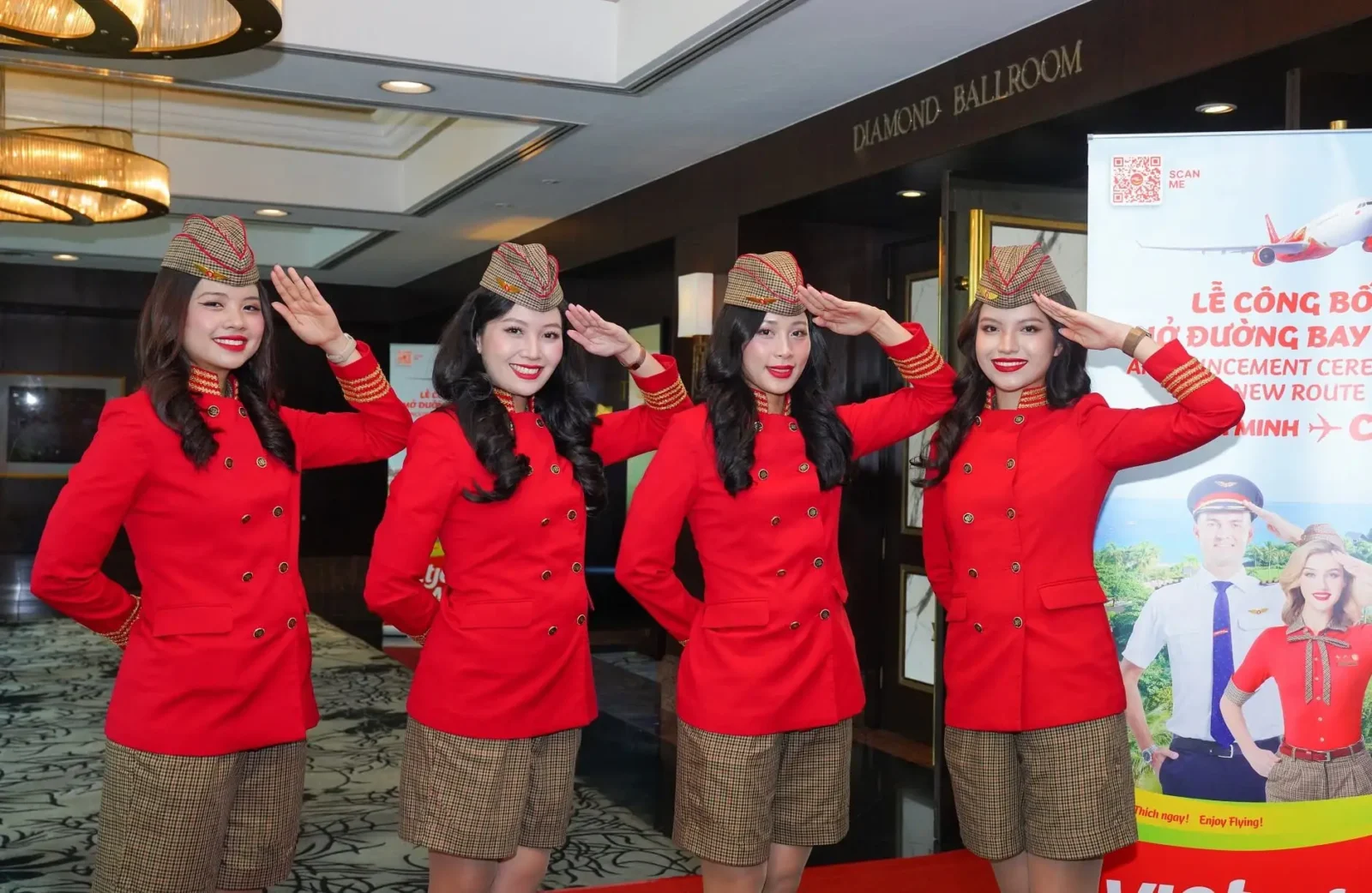 Four Vietjet Air cabin crew members in red uniforms and patterned caps saluting at a new route announcement ceremony, standing in front of a Vietjet promotional banner inside a hotel ballroom.
