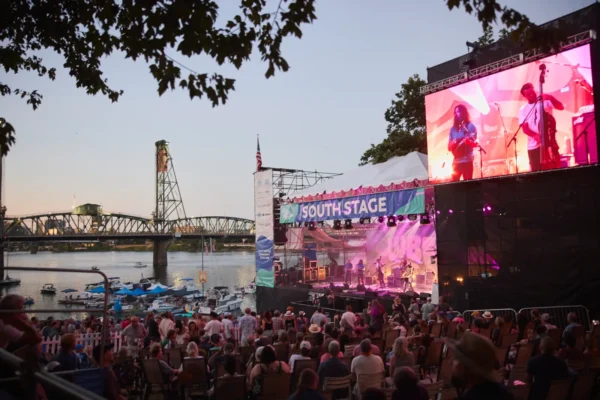 Evening crowd seated and standing before the South Stage at Waterfront Blues Festival, with a band performing under pink and purple lighting, a large video screen, boats on the Willamette River and the Hawthorne Bridge in the background