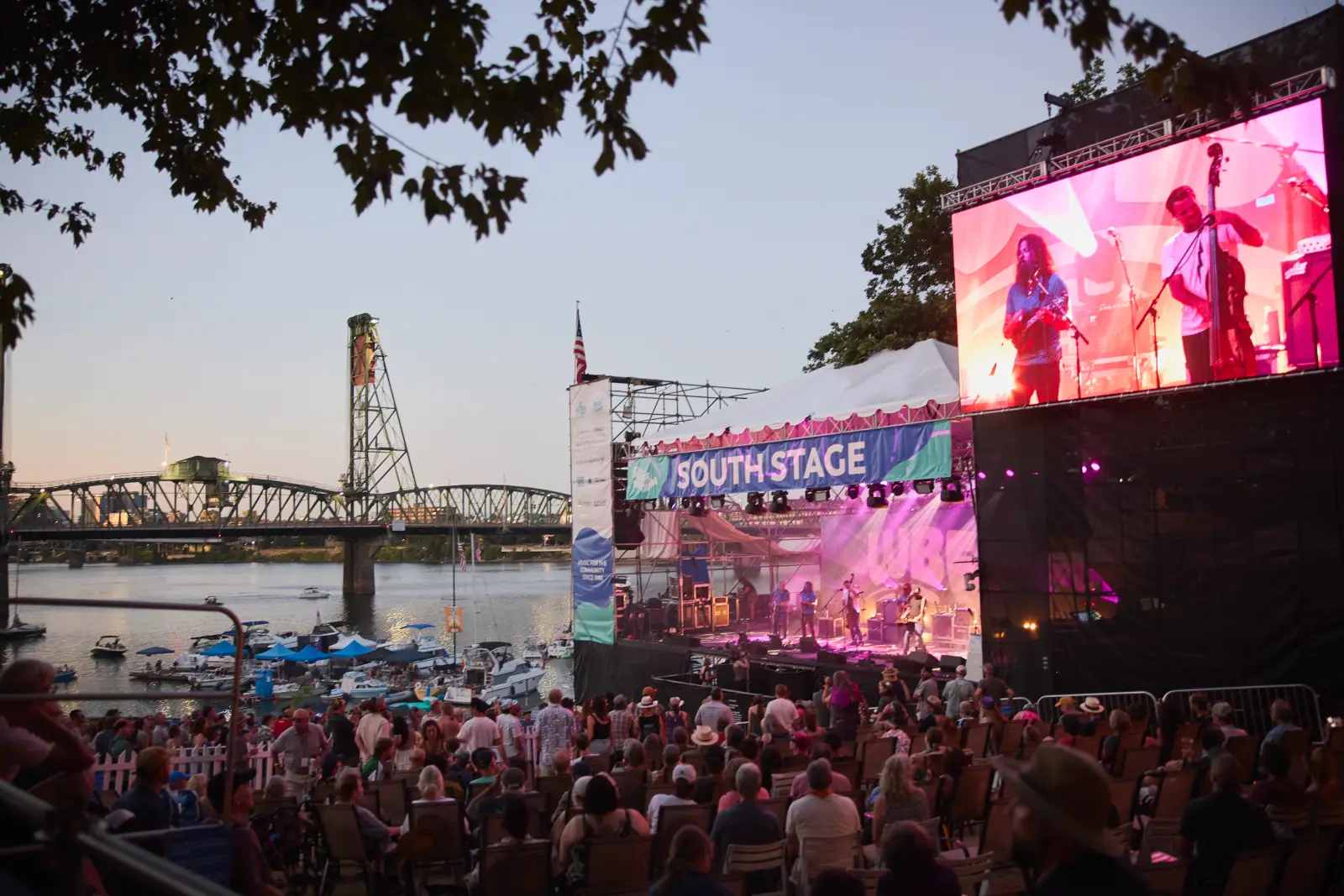 Evening crowd seated and standing before the South Stage at Waterfront Blues Festival, with a band performing under pink and purple lighting, a large video screen, boats on the Willamette River and the Hawthorne Bridge in the background