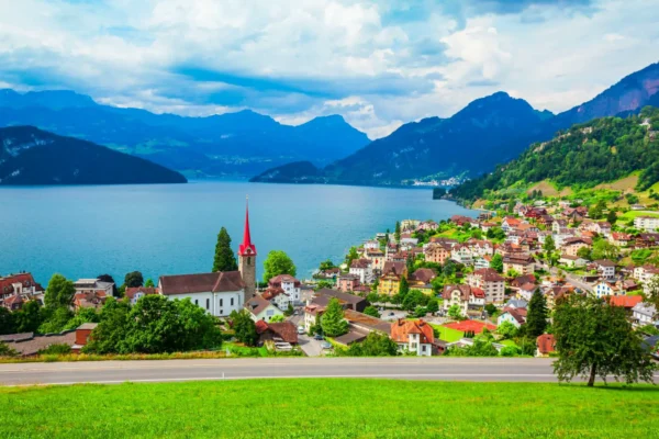 Home 12 Panoramic view of Weggis on Lake Lucerne with a church, lakeside village, and surrounding Swiss Alps under a dramatic sky.
