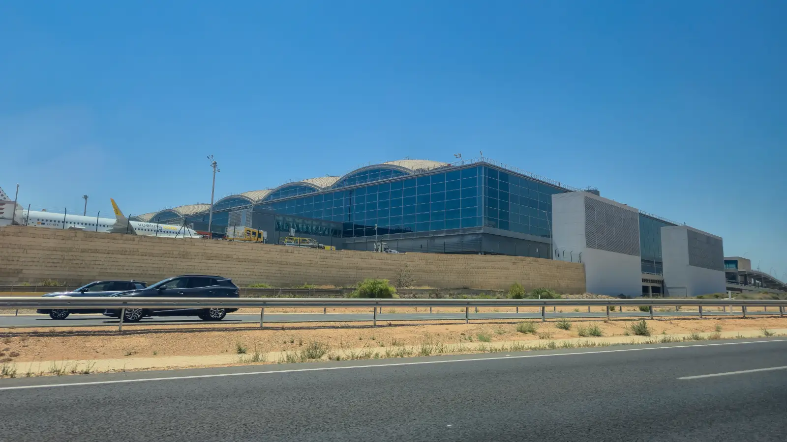 Exterior view of Alicante-Elche Miguel Hernández Airport's terminal, showing its curved roof design and glass facade under a clear sky