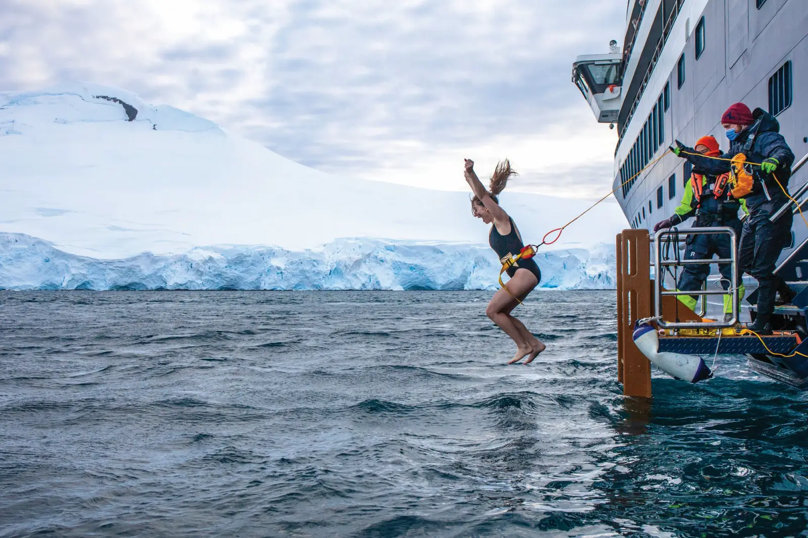 Atlas Ocean Voyages Expands Fly-Cruise Antarctica Expeditions For 2027-2028 Season 3 A guest secured by a harness leaps into the icy waters off Antarctica during the complimentary Polar Plunge, with crew members assisting from the ship's platform and glaciers visible in the background.