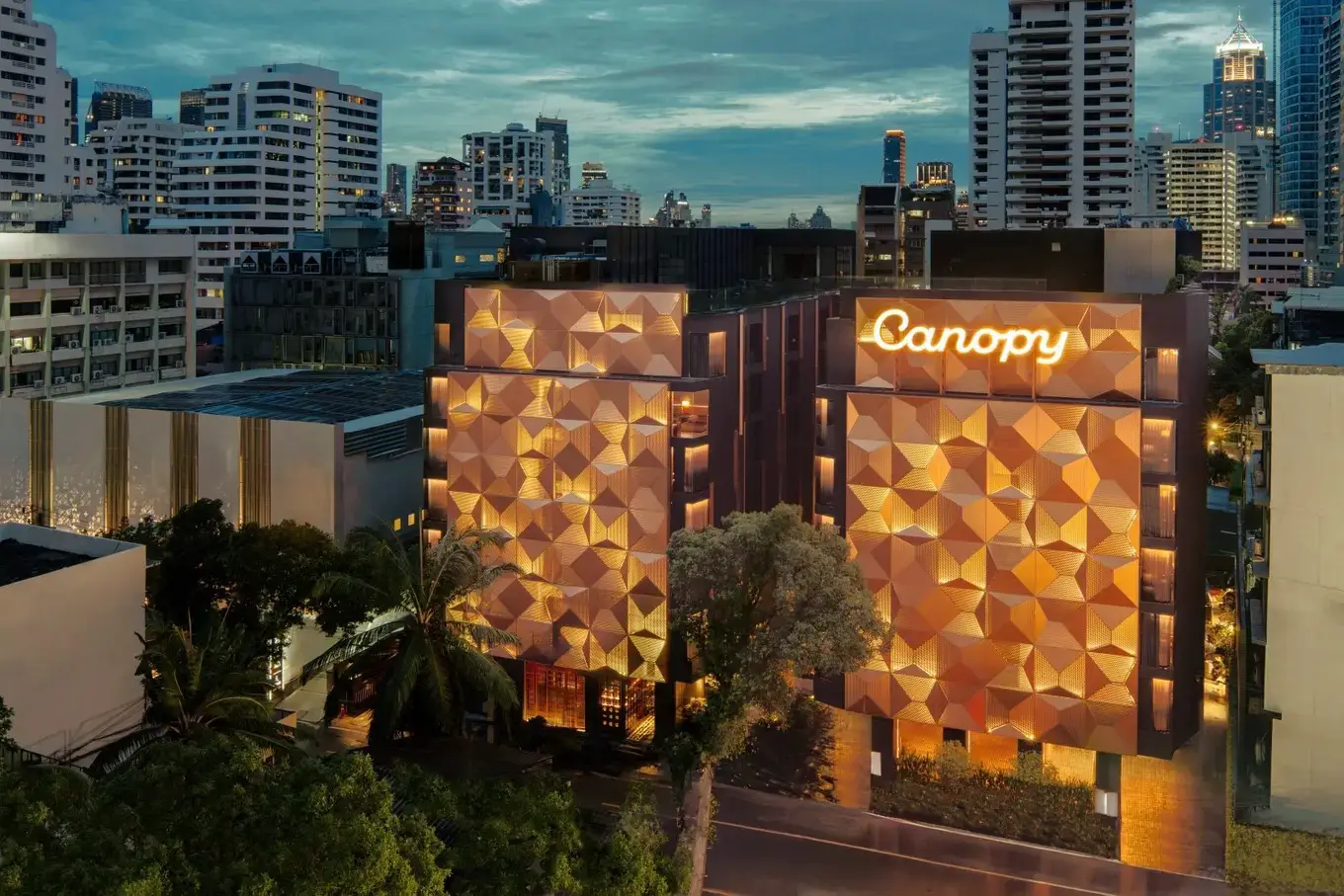 Canopy by Hilton Bangkok hotel exterior at dusk with illuminated geometric facade and city skyline.