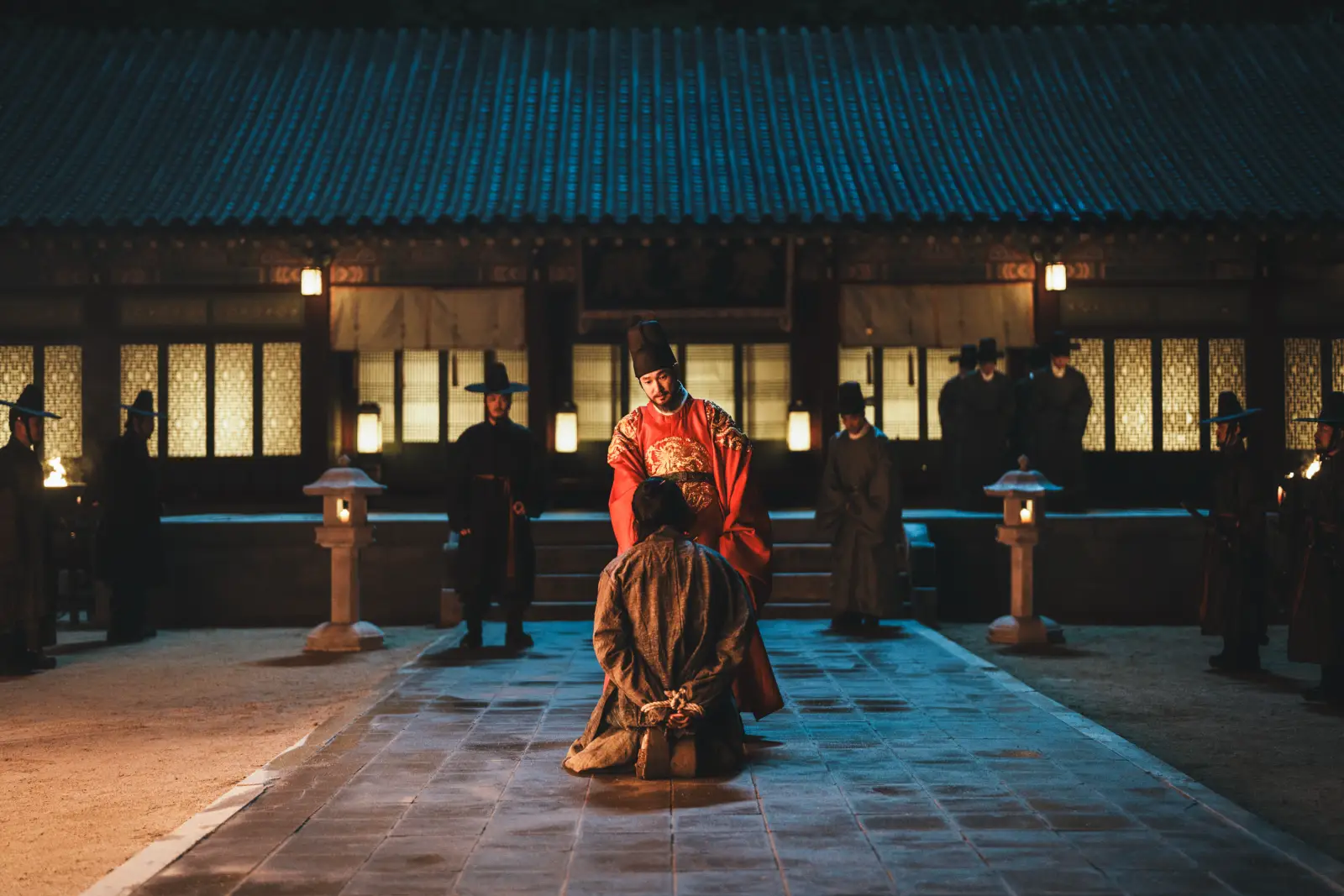 A royal figure confronts a kneeling prisoner in a torchlit palace courtyard in The East Palace.