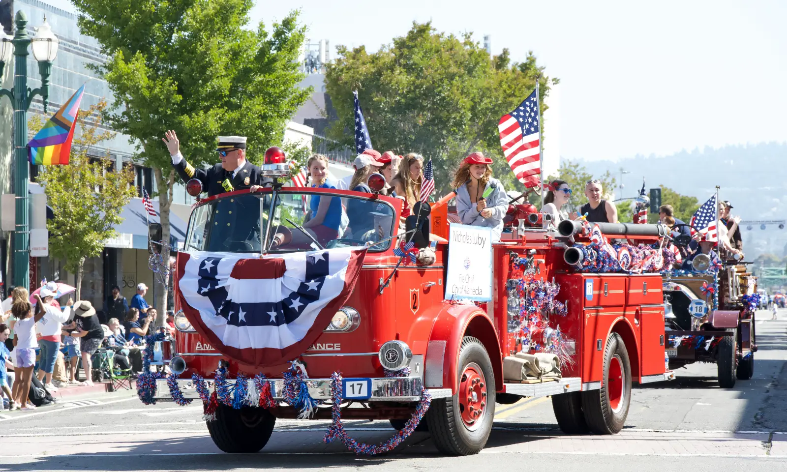 Vintage red fire truck decorated with American flags and bunting leads a Fourth of July parade past cheering crowds