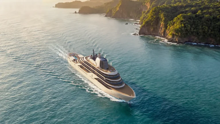 A Four Seasons luxury yacht cruises along a tropical coastline with lush green cliffs and calm blue waters at sunset.
