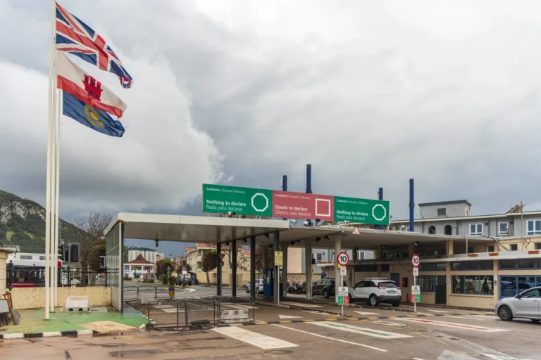 Cars pass through the Gibraltar border customs checkpoint beneath British and Gibraltar flags.
