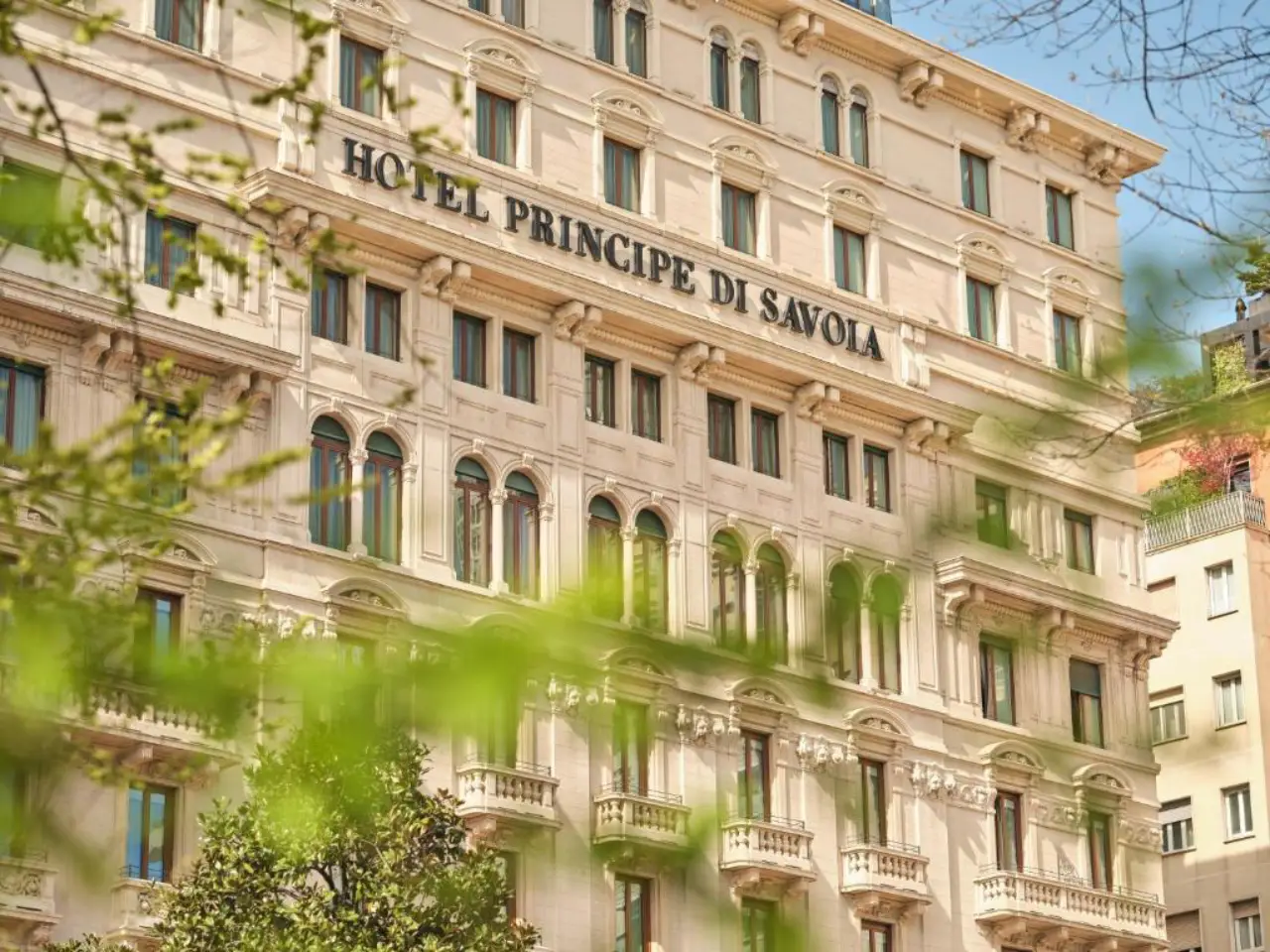 Historic façade of Hotel Principe di Savoia in Milan, Italy, viewed through spring greenery