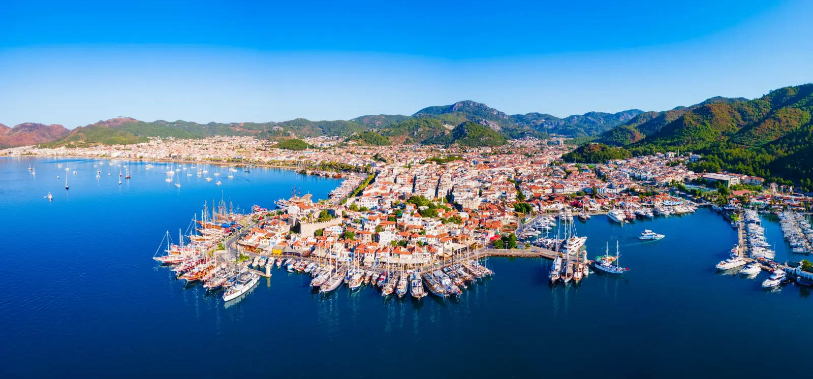 Aerial panoramic view of Marmaris Marina and waterfront with yachts, the historic castle and surrounding mountains in Muğla, Türkiye.