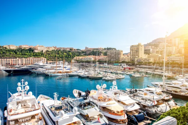 Home 5 Luxury yachts moored at Port Hercule marina in Monaco with the city skyline and Mediterranean coastline under a sunny blue sky.
