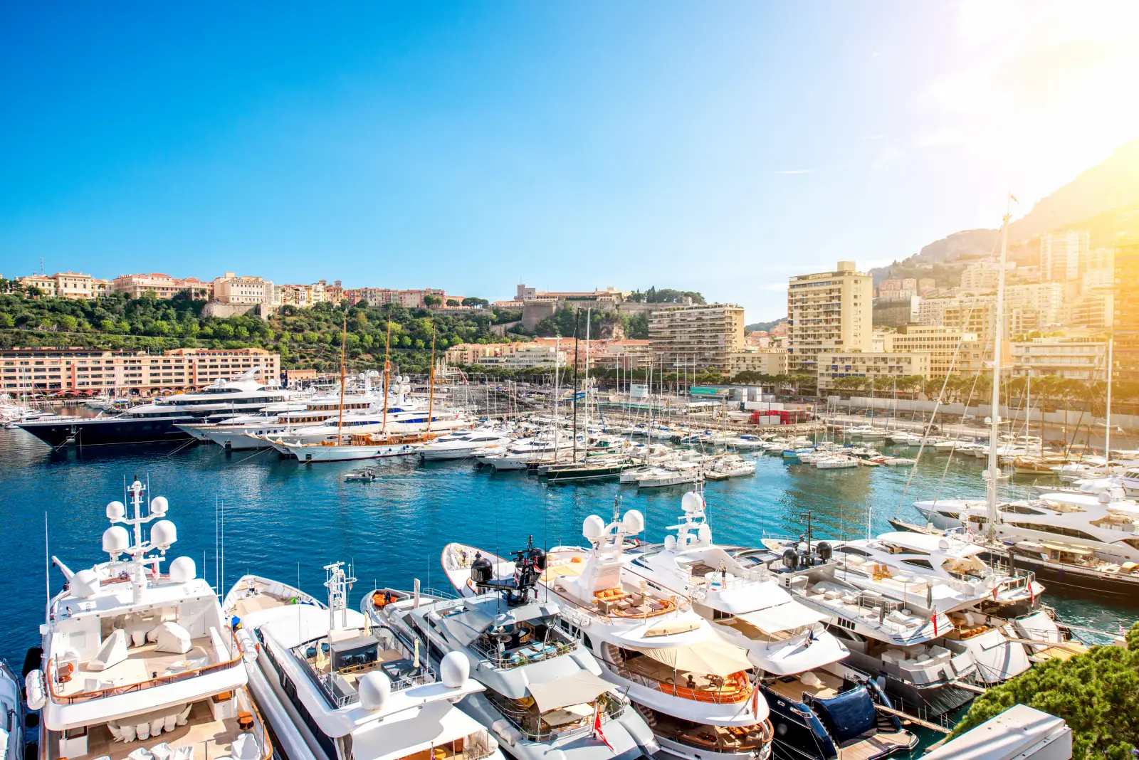 Luxury yachts moored at Port Hercule marina in Monaco with the city skyline and Mediterranean coastline under a sunny blue sky.