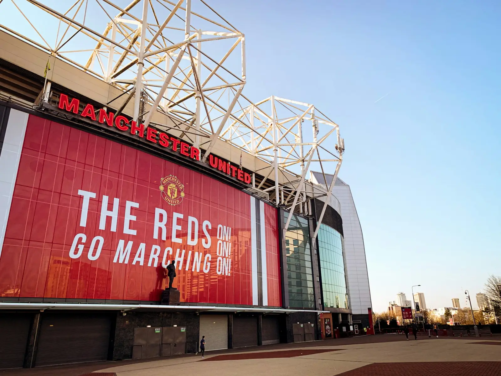 What To Expect From Traveling To Manchester 3 Exterior view of Old Trafford, home of Manchester United, featuring the stadium facade and iconic club signage under a clear blue sky.