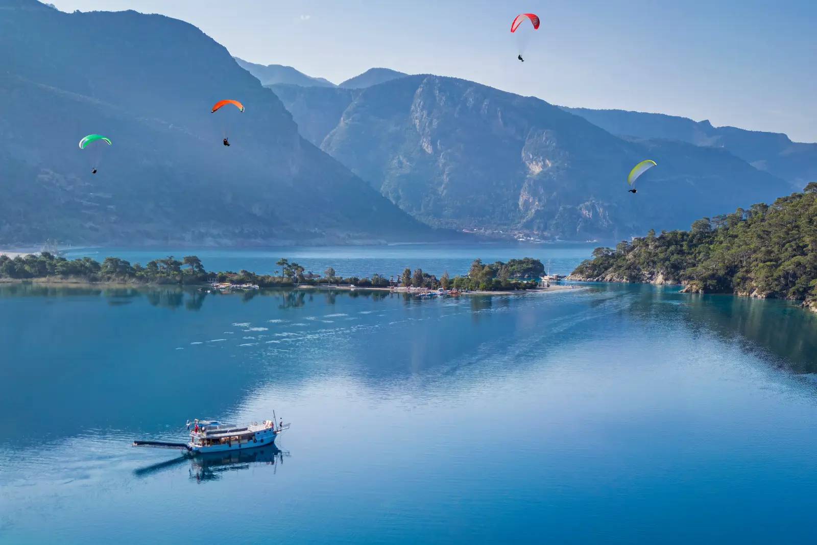 Ölüdeniz Paragliding Pilots Halt Flights Over Rising Costs 3 Paragliders soaring above Ölüdeniz Beach and the Blue Lagoon in Fethiye, Türkiye, with turquoise waters, a sightseeing boat and surrounding mountains