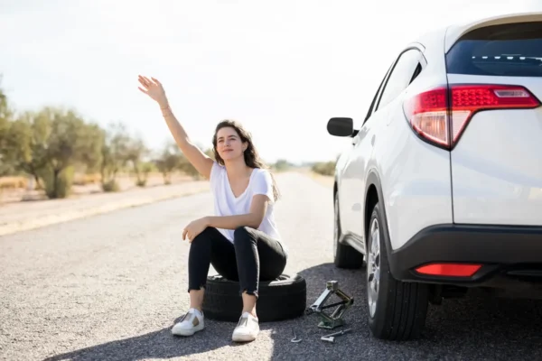 Home 10 A driver sits beside a white SUV with a flat tire, waving for roadside assistance after a breakdown on a rural road.