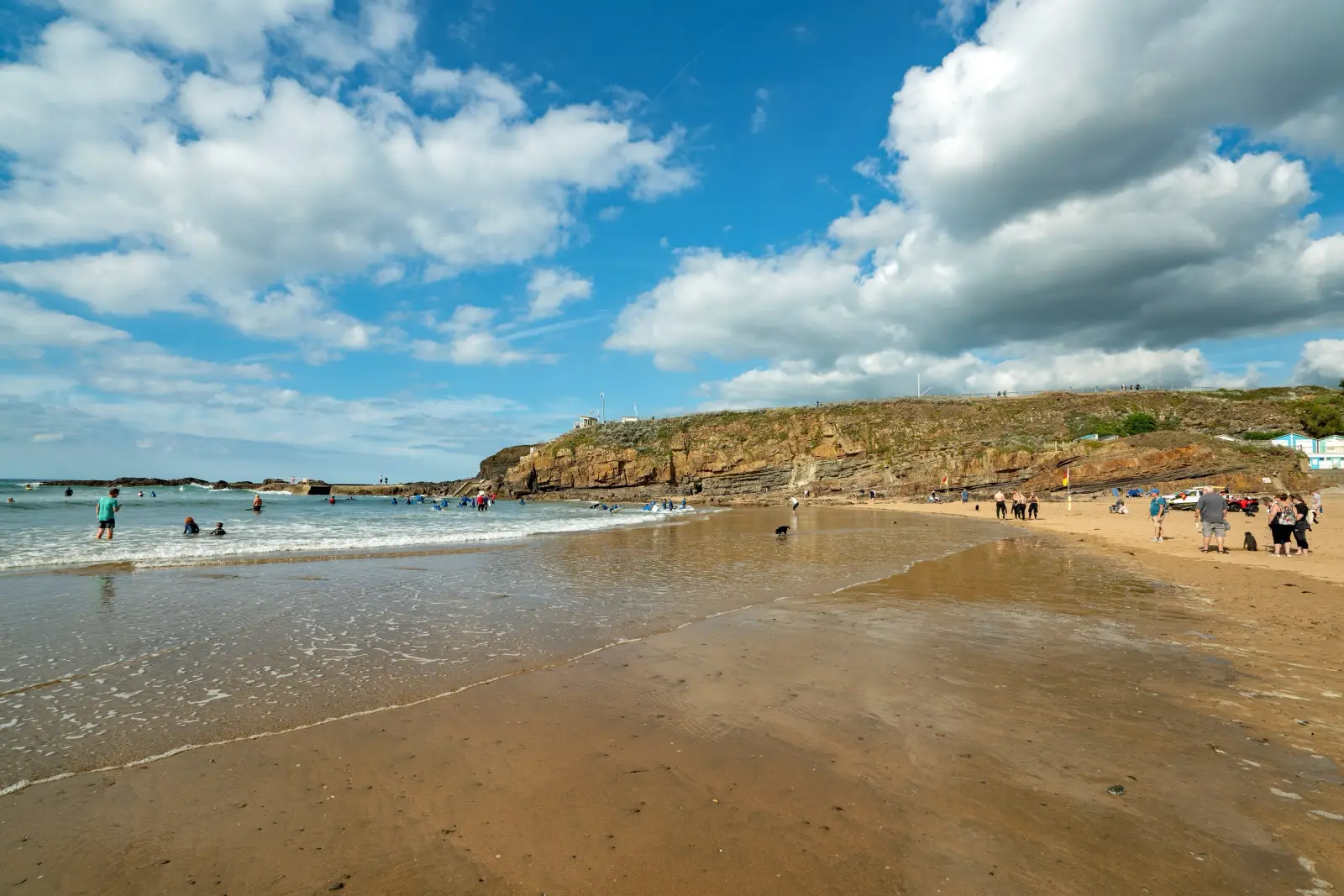Visitors enjoy Summerleaze Beach in Bude, Cornwall, on a sunny day with surfers, sandy shoreline and dramatic coastal cliffs