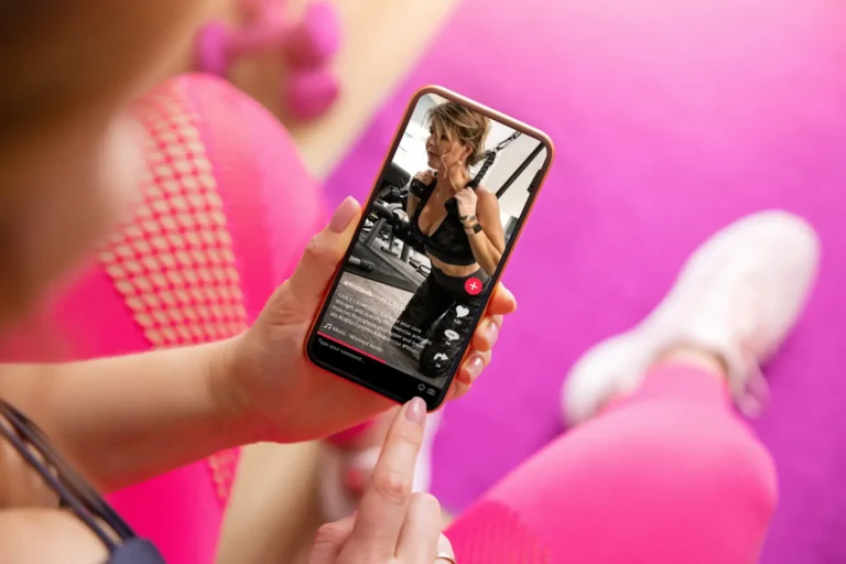Person holding a smartphone displaying a TikTok fitness video while sitting on a yoga mat with workout equipment nearby.