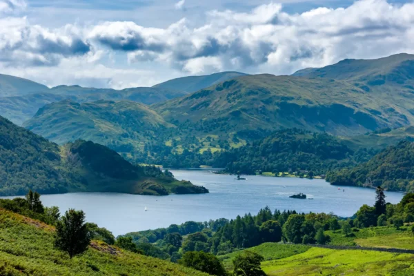 Panoramic summer view of Ullswater from Gowbarrow Fell in England's Lake District, with green hills, a blue lake, and scattered clouds.