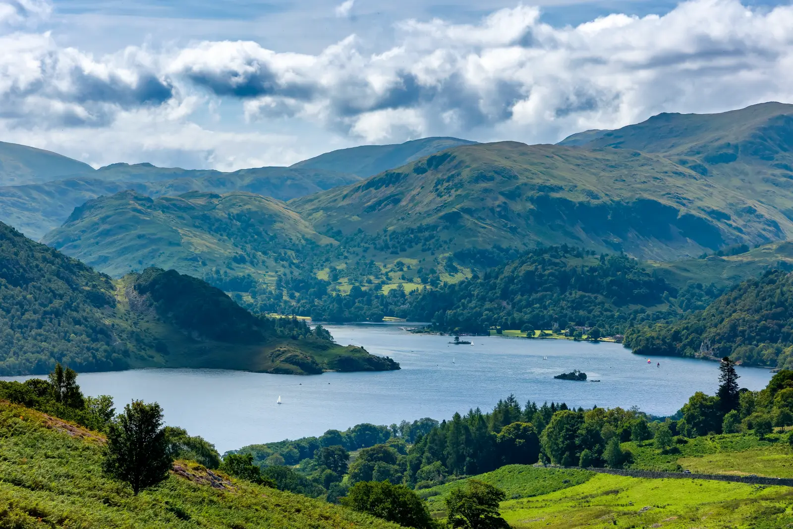 Cumbria Travel Pass Opens Lake District To Unlimited Travel 4 Panoramic summer view of Ullswater from Gowbarrow Fell in England's Lake District, with green hills, a blue lake, and scattered clouds.